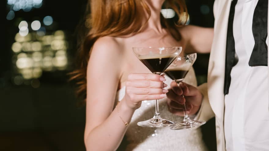 Close up of woman in white strapless wedding dress and man wearing a white dining coat, white button-down shirt and an un-done black bow tie clinking together cocktail glasses