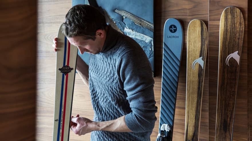Man wearing knit sweater leans over ski against wall, more skis on wall