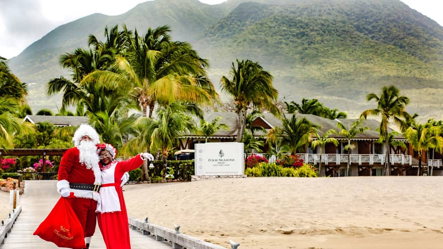 Couple dressed up as Santa and Mrs. Claus stand in front of Resort sign, mountains and palm trees visible in background
