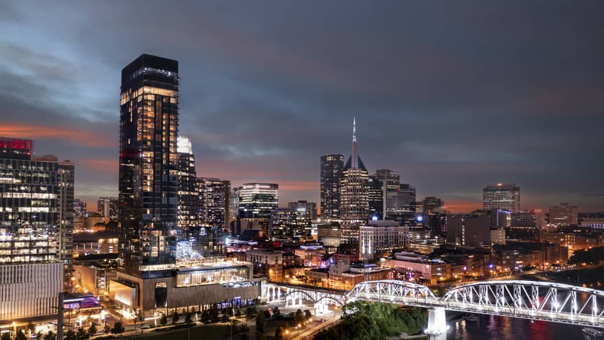 Nighttime view of downtown Nashville with illuminated skyscrapers, a lit-up bridge, and a vibrant cityscape under a dusky sky.