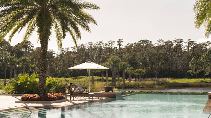 Outdoor swimming pool where woman lounges on patio chair under palm tree