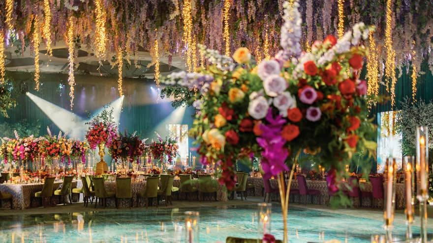 Ballroom decorated with large bright yellow, pink, red and white floral arrangements and purple wisteria hanging from the ceiling over a blue dancefloor