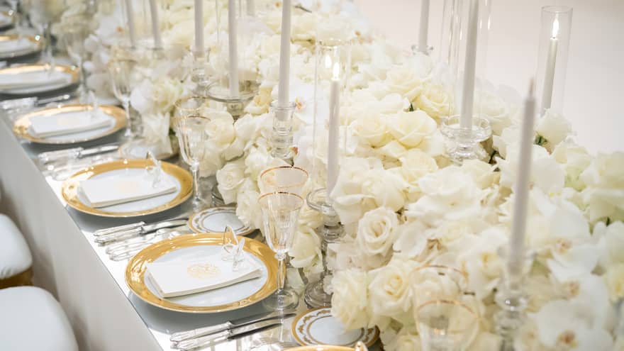 Table filled with white flowers and candles in the centre with a row of white, gold-rimmed plates on either side