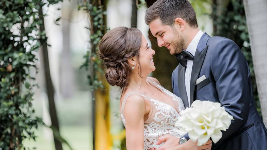 Smiling groom and bride with white wedding dress, bouquet embrace