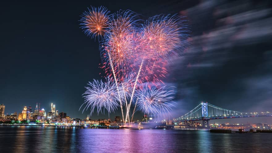 Purple, pink and blue fireworks exploding in the night sky above a city and a large metal bridge.