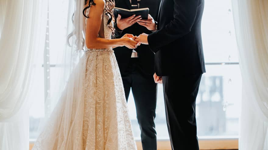 Bride and groom hold hands as wedding officiant reads from book by sunny window