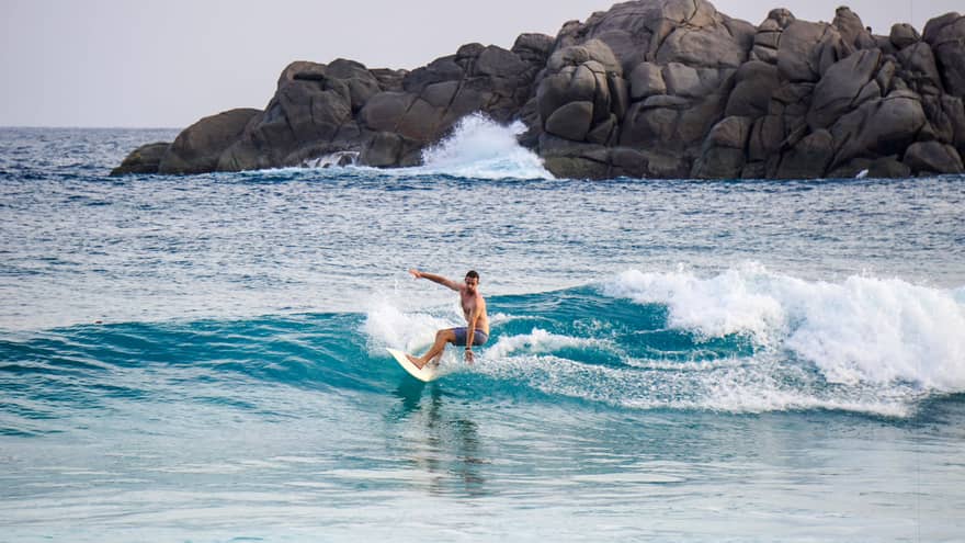 Man on surfboard rides on small blue wave, large rocks in background