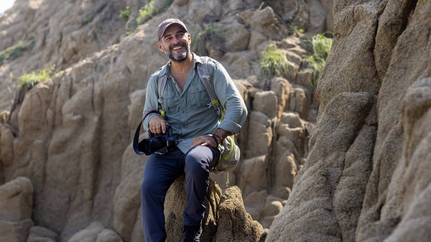 A photographer wearing a backpack and holding a camera in one hand sits atop a rock formation, smiling into the distance.