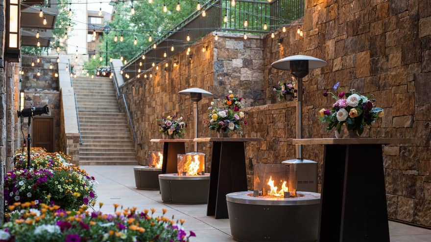 Outdoor event space with stone walls, string lights, floral arrangements on tall tables and firepits creating a warm ambience 