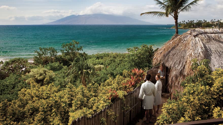 Couple in white bathrobes follow spa attendant into thatched-roof hut on path looking out at ocean