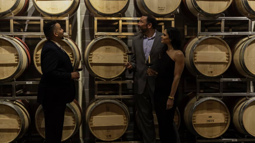 A couple in evening wear hold glasses of red wine and listen to a sommelier while looking at a rack of wine barrels.
