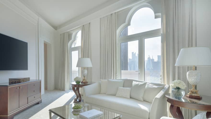 A light-filled living room with white sofa, white arm chair, tan carpet, glass coffee table, lamps