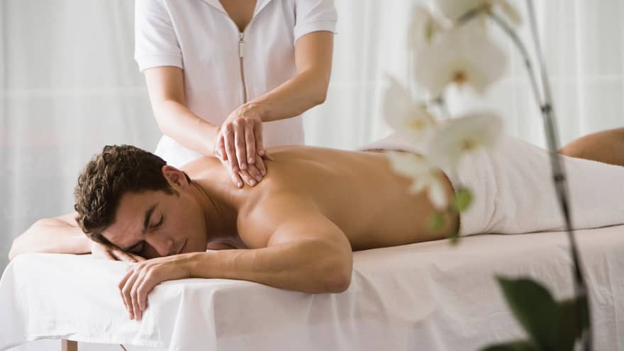 Man lays on white table with eyes closed as masseuse massages his bare shoulders