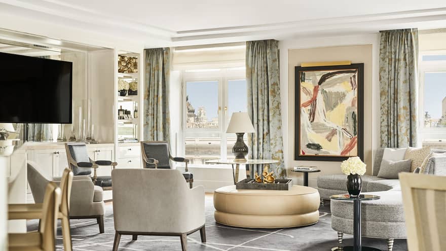 Hotel suite with abstract artwork, TV, grey armchairs and sofa, round beige ottoman, modern decor, glass tables and floor-to-ceiling windows with city views