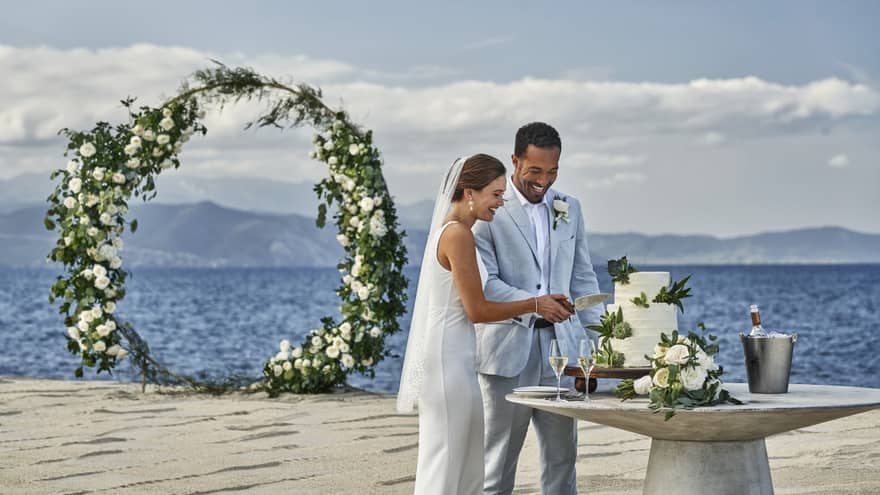 A bride and groom joyfully cut their wedding cake on a beautiful beach, surrounded by ocean waves.