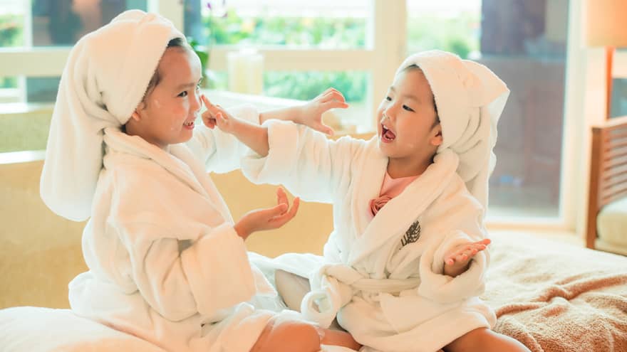 Two laughing young children in white bathrobes with towels around hair put lotion on each other's faces 
