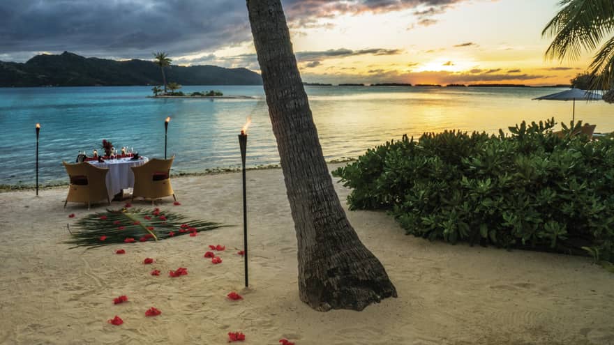 Rose petals and torches lead to a candlelit dinner table on the white sand beach by shore  at sunset