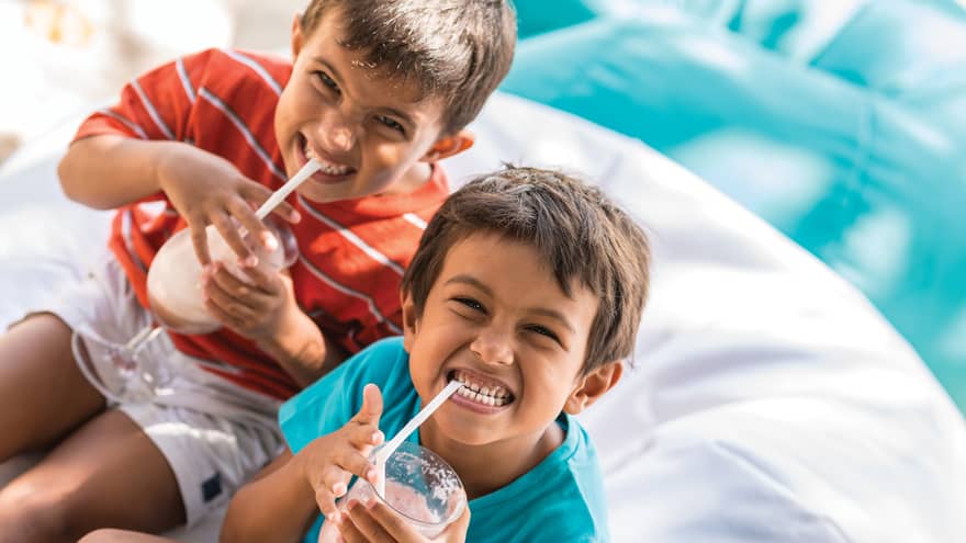 Two boys smile really big smiles as they sip on a virgin piña colada on a bean bag cushion