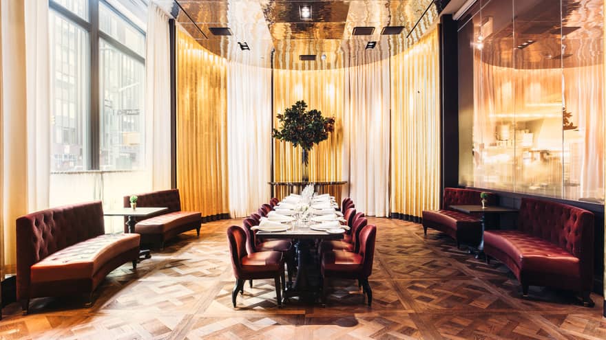 Sofas, long dining table with red velvet chairs under tall brass ceiling and floor-to-ceiling mirror