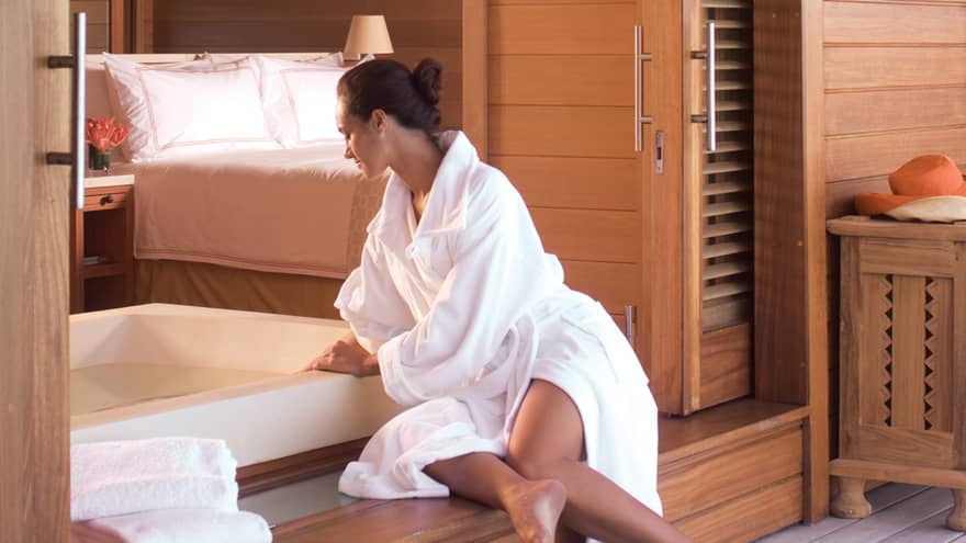 Woman in white bathrobe prepares bath inside bungalow, with bed in background
