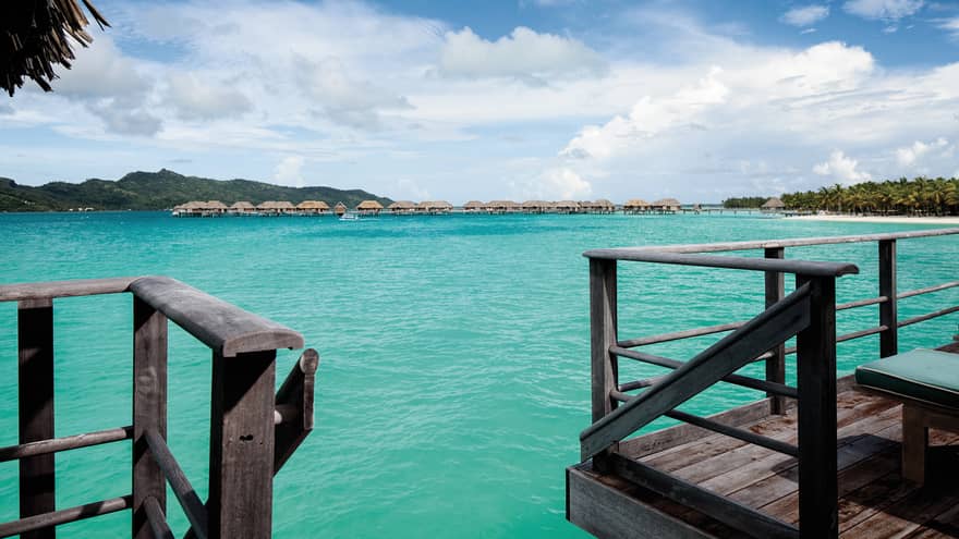 View from steps of overwater bungalow deck leading to blue lagoon, more bungalows, mountains in far view