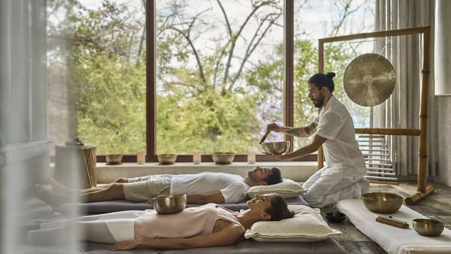 A man using sound therapy while a couple is lying down near a window looking out at trees.