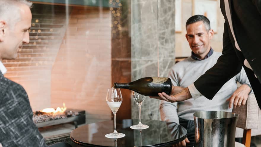 Two men being poured a glass of wine while sitting next to a fire.