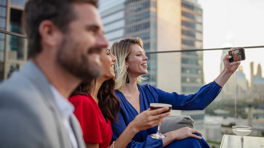 A man and two women sit and socialize on outdoor terrace with city building in the background