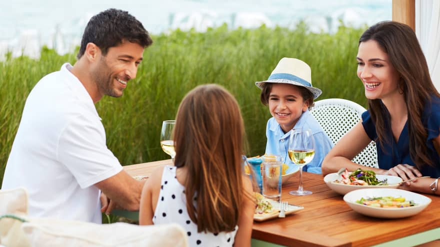 A family of four smiles as they enjoy a meal beachside on an outdoor patio