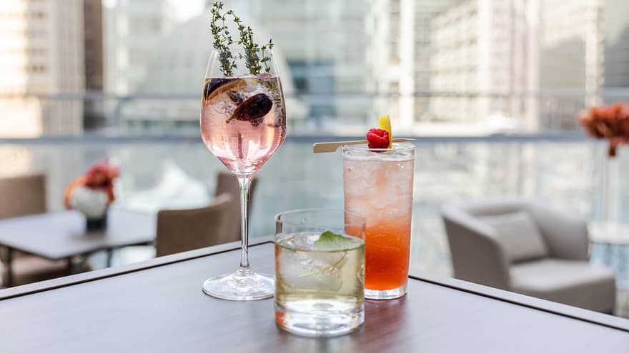 A pink, red and clear cocktail sitting on the corner of a table on a rooftop outside, tall buildings can be seen in the background.