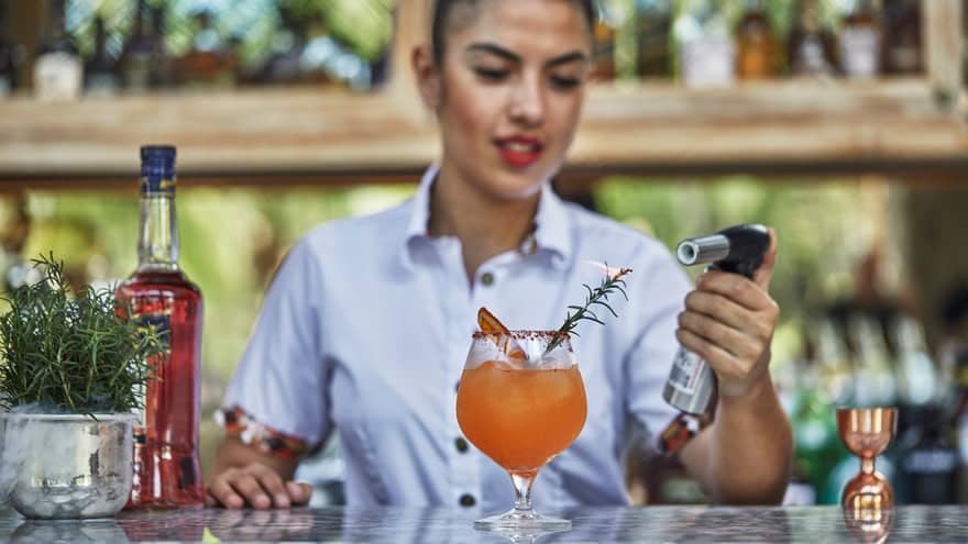 A female bartender mixes an orange drink, and is using a blow torch on the top.