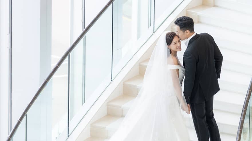 A bride and groom walking up stairs near large windows.