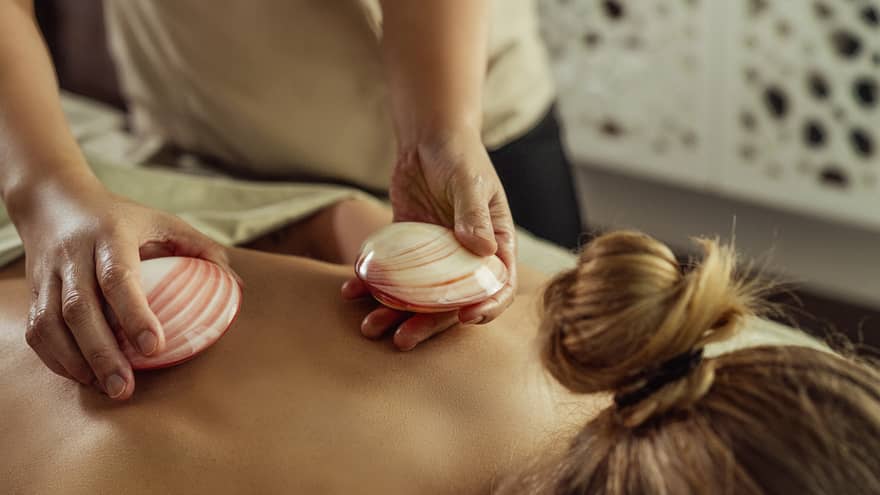 Close up of masseuse's hands placing red-and-white shells on the bare back of a person lying face down