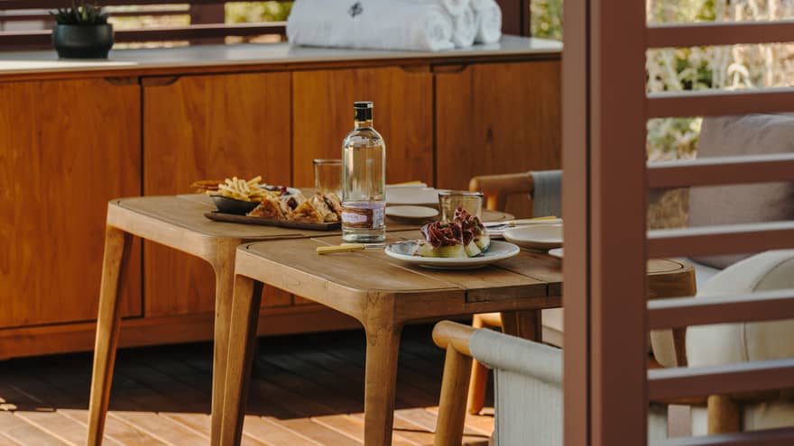 A private pool cabana with small tables, comfortable seating, a cabinet with rolled-up towels and slatted walls admitting filtered light sunlight.
