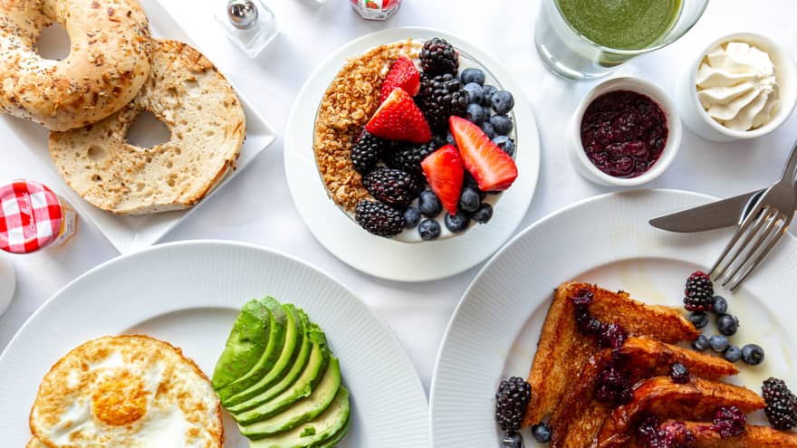 Overhead view of French toast, fried eggs, sliced avocado, granola and fruit, a bagel and various jams spread on a white table