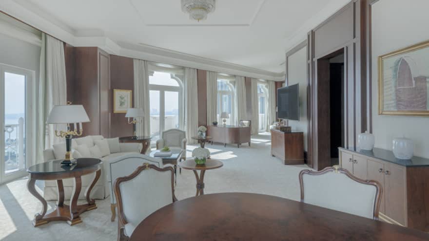 Large living area of hotel suite, with dark wooden dining table, white sofa and many windows lining the wall