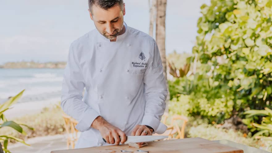 A male chef thinly slices seafood on wooden cutting board, beach in the background