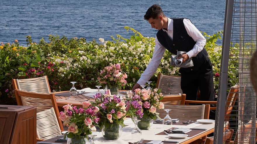 Waiter sets outdoor table with pink floral centrepieces, ocean and mountains in the background