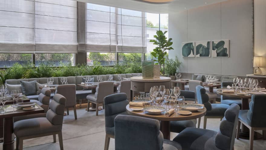 Modern restaurant dining room with beige banquette seating against tall windows and a mix of round and square dining tables