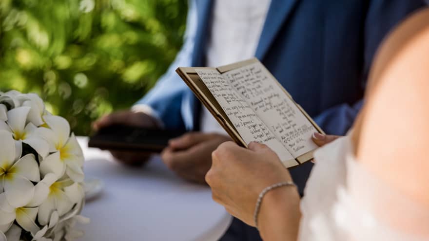 Bride holds written vows beside groom holding vows atop white table