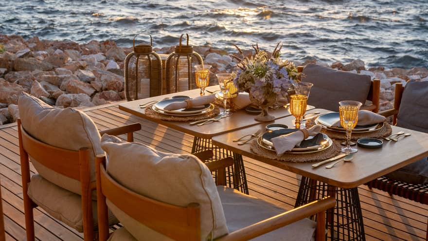 A table laid with sparkling glasses and a bouquet of flowers set on a deck on the rocky shore, the sunlit waves metres away.