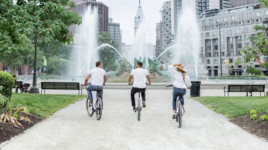 Three guests riding bikes towards a large fountain with the Philadelphia skyline in the distance