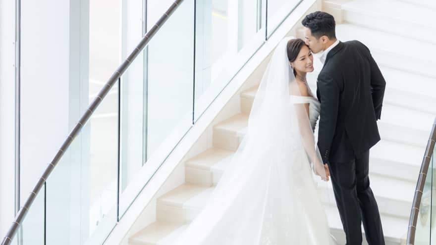 A bride and groom walking up stairs near large windows.