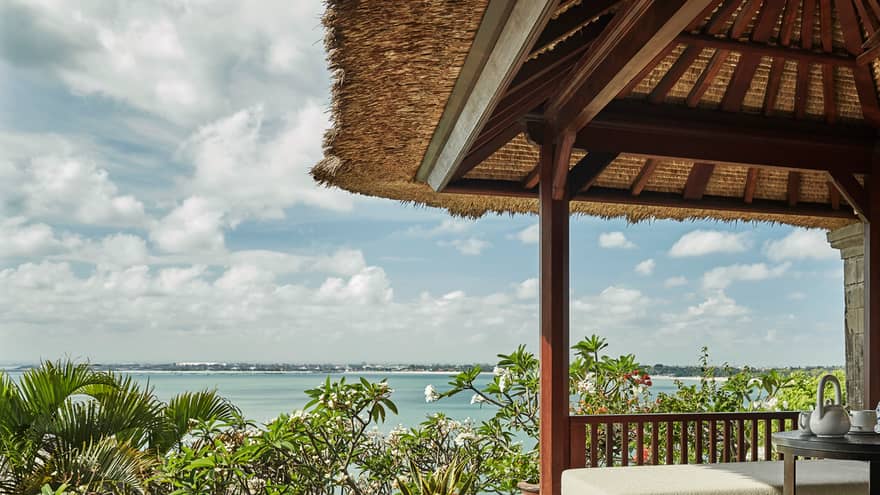 Close-up of Deluxe Villa patio corner with swimming pool, plant, white bench under thatched roof, ocean view