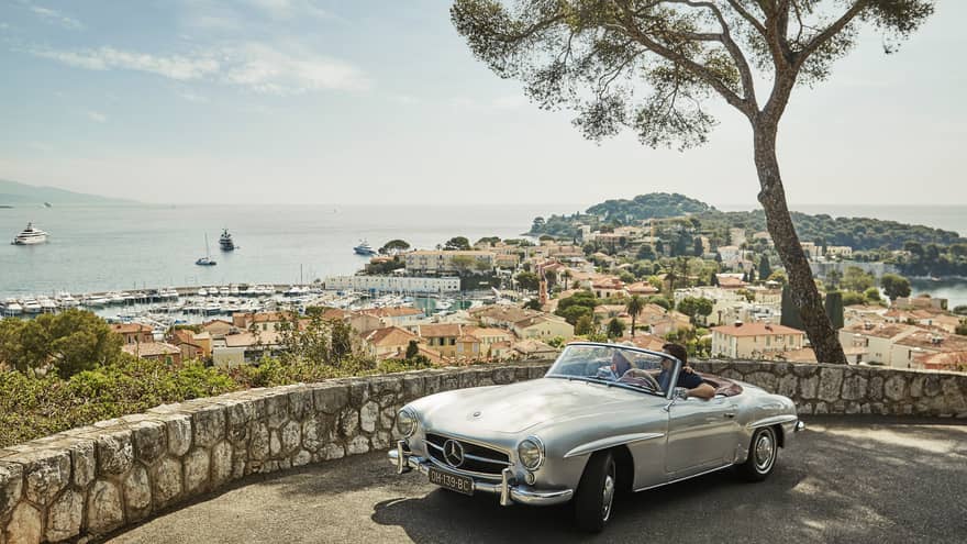Couple in luxury vintage white car parked under tree by stone wall overlooking red roofs, water