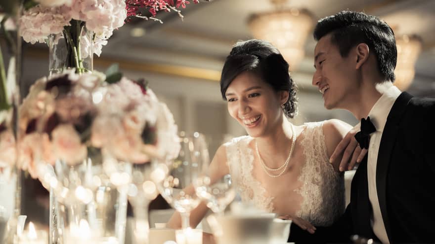 Smiling bride and groom at wedding dining table with glowing candles, pink roses