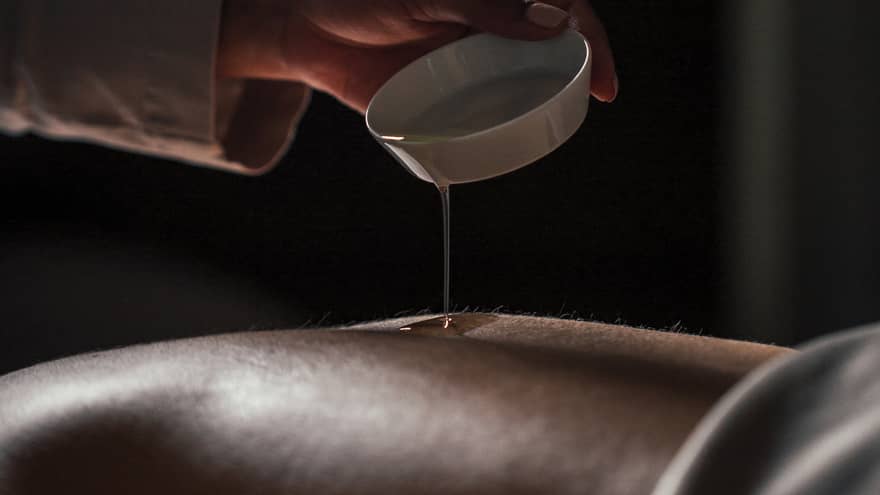 Spa staff pours oil over person's bare back in dark room