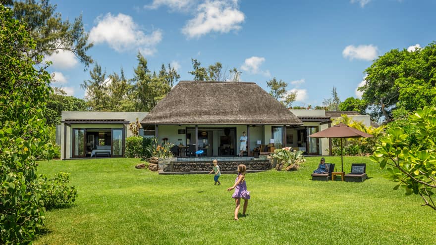 Young children play on green lawn in front of bungalow as dad watches from covered patio