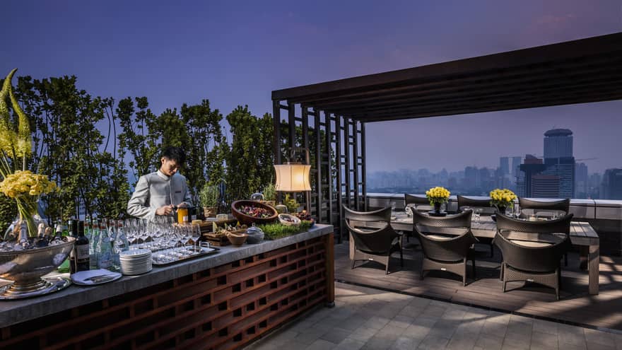 Bartender prepares drinks, cheeses, salads at long Imperial Suite rooftop bar by private dining table at night