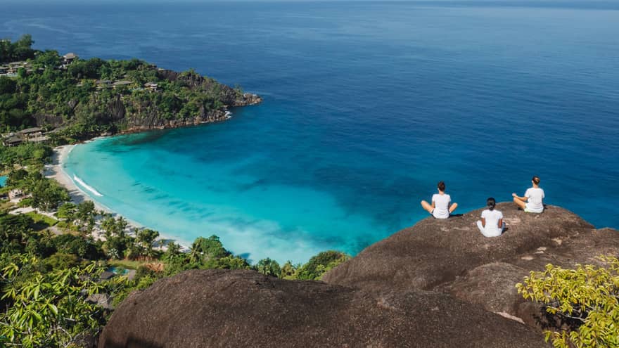 Three people sit cross-legged in meditation overlooking the ocean.